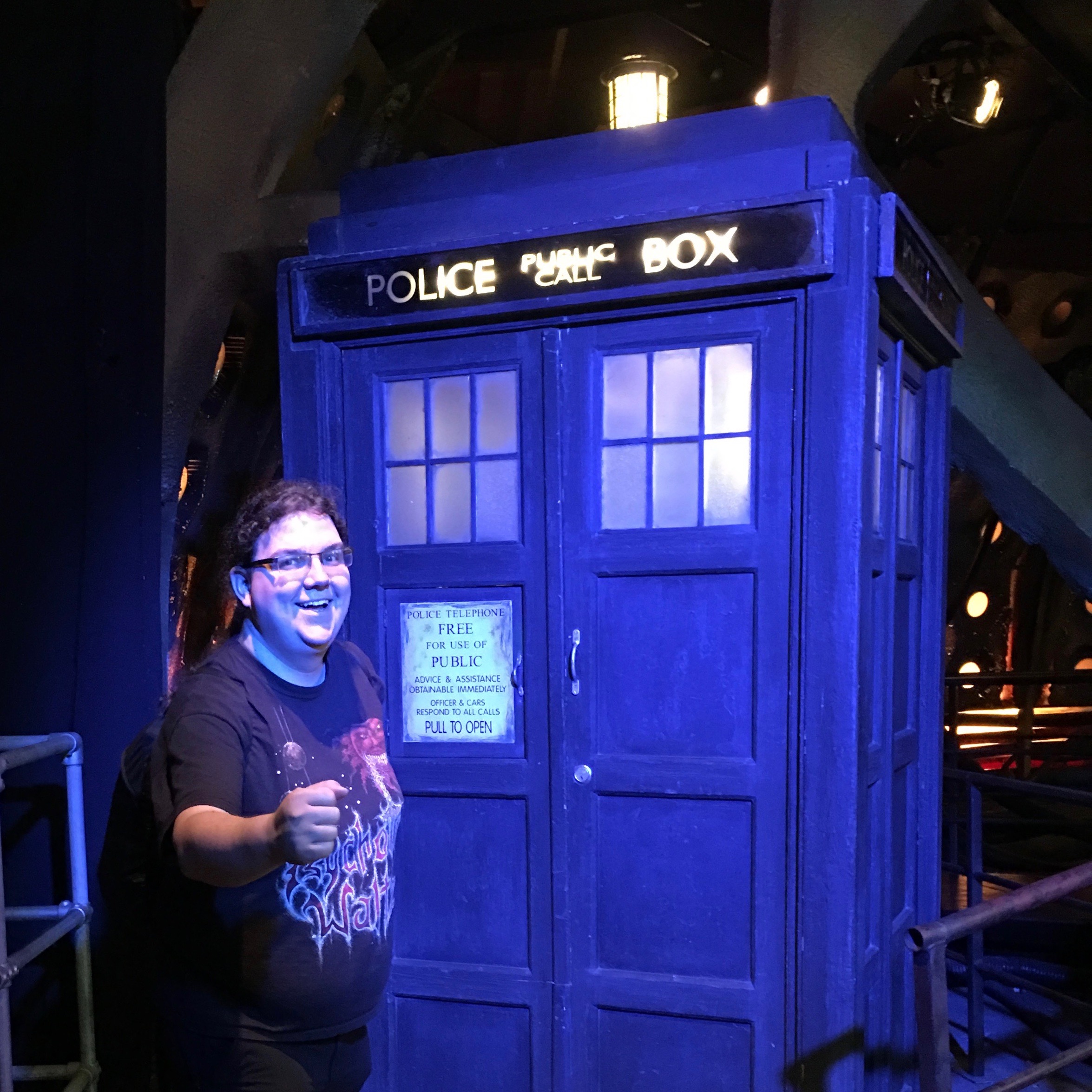 Eric Eggert, a long-haired overweight white person wearing a Metal t-shirt stands in front of an original, used on set TARDIS prop from Doctor Who in the Doctor Who Experience in Cardiff.